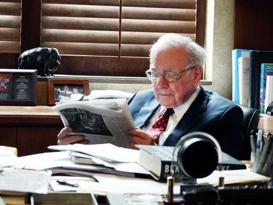 Warren Buffett ngồi ở bàn làm việc, đeo kính, đọc một tờ báo gấp đôi, trước mặt là chồng giấy và sách